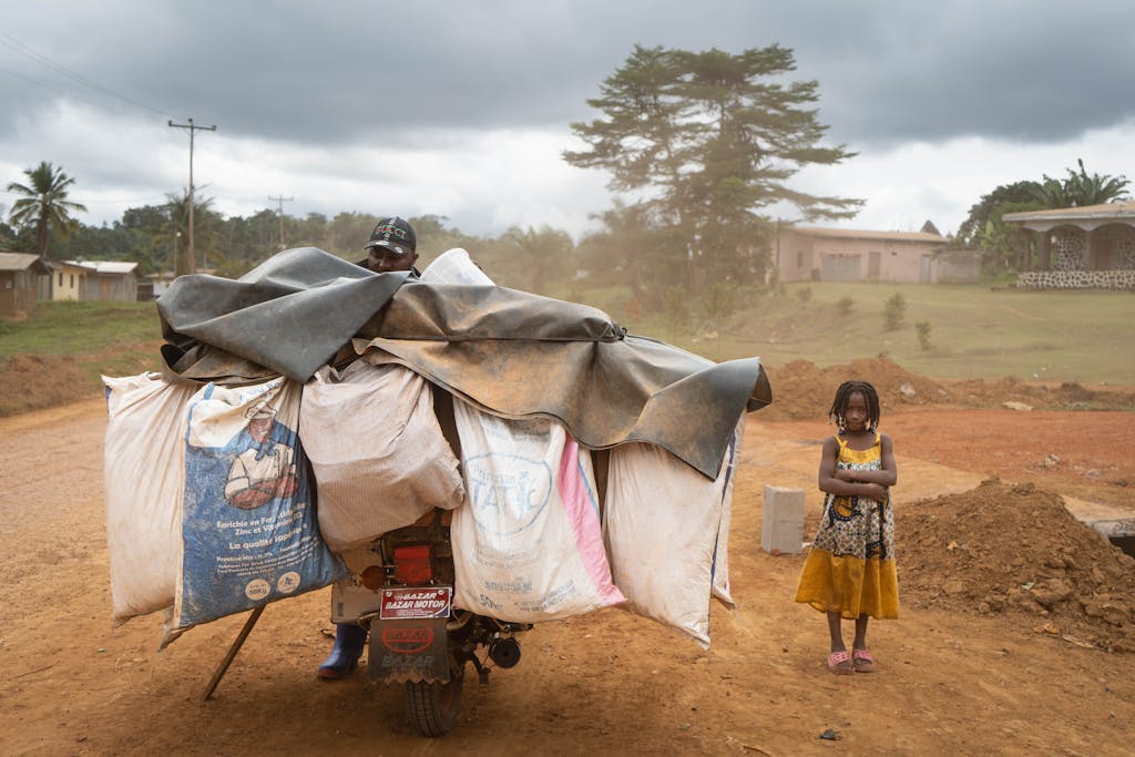 A young girl stands beside a loaded motorcycle on a rural Cameroonian road.