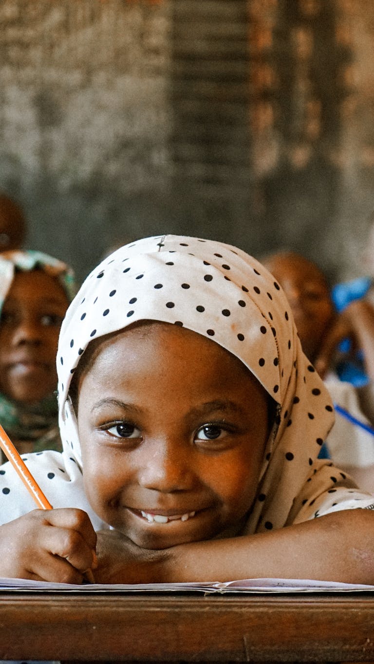 Young girl smiles while writing in a lively classroom setting, capturing joyful learning.